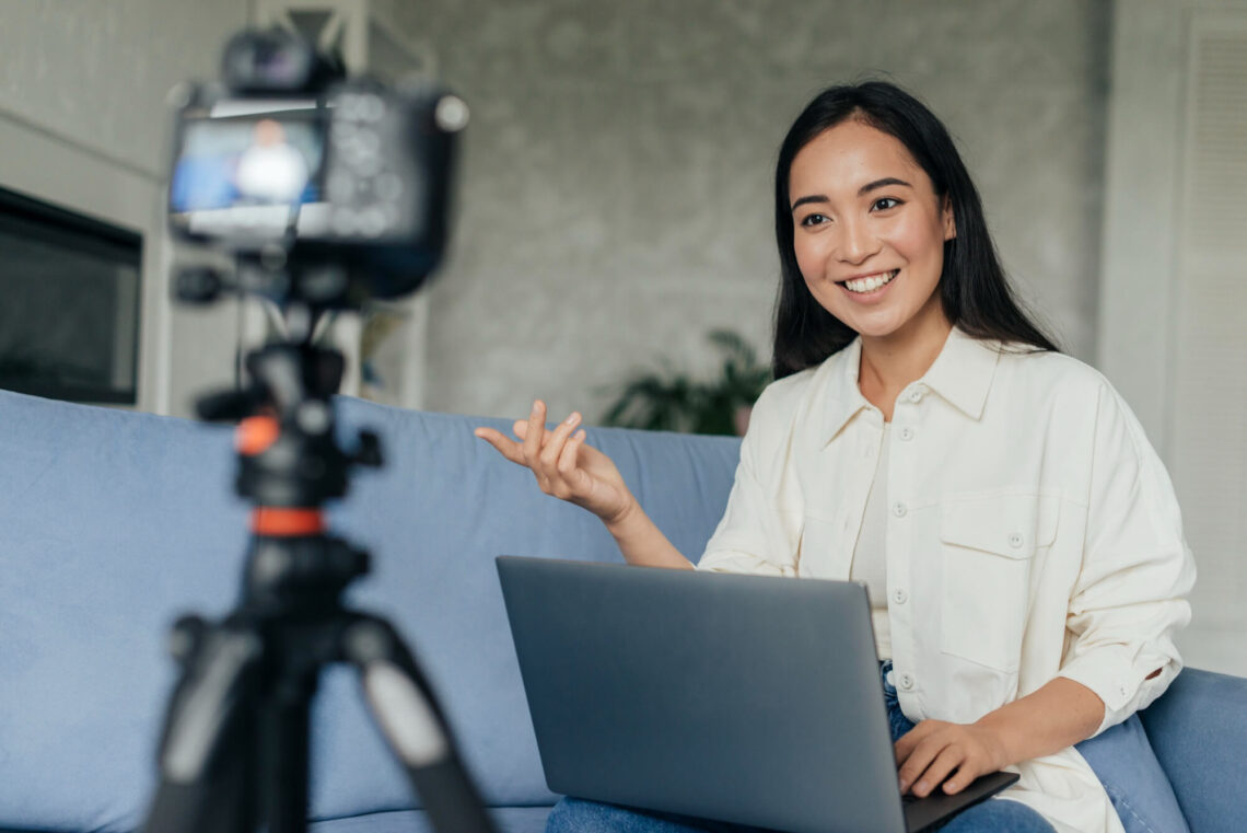 Mulher sentada no sofá com notebook no colo e olhando para a câmera para fazer uma transmissão ao vivo.