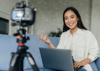 Mulher sentada no sofá com notebook no colo e olhando para a câmera para fazer uma transmissão ao vivo.